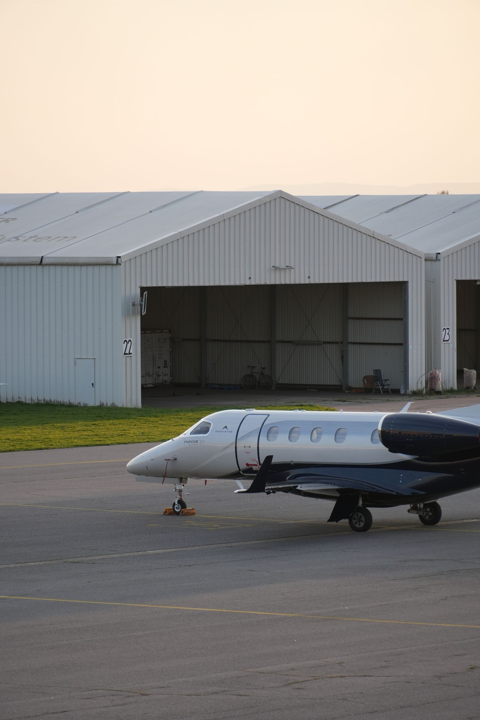 An Embraer Phenom 300 business jet parked on a ramp in front of a row of white hangars at golden hour