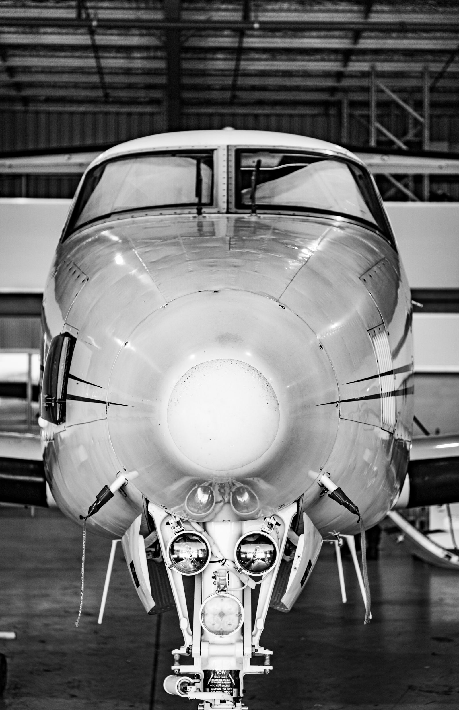 Black-and-white head-on close-up of a business jet's nose, landing light and nose gear, parked inside a hangar