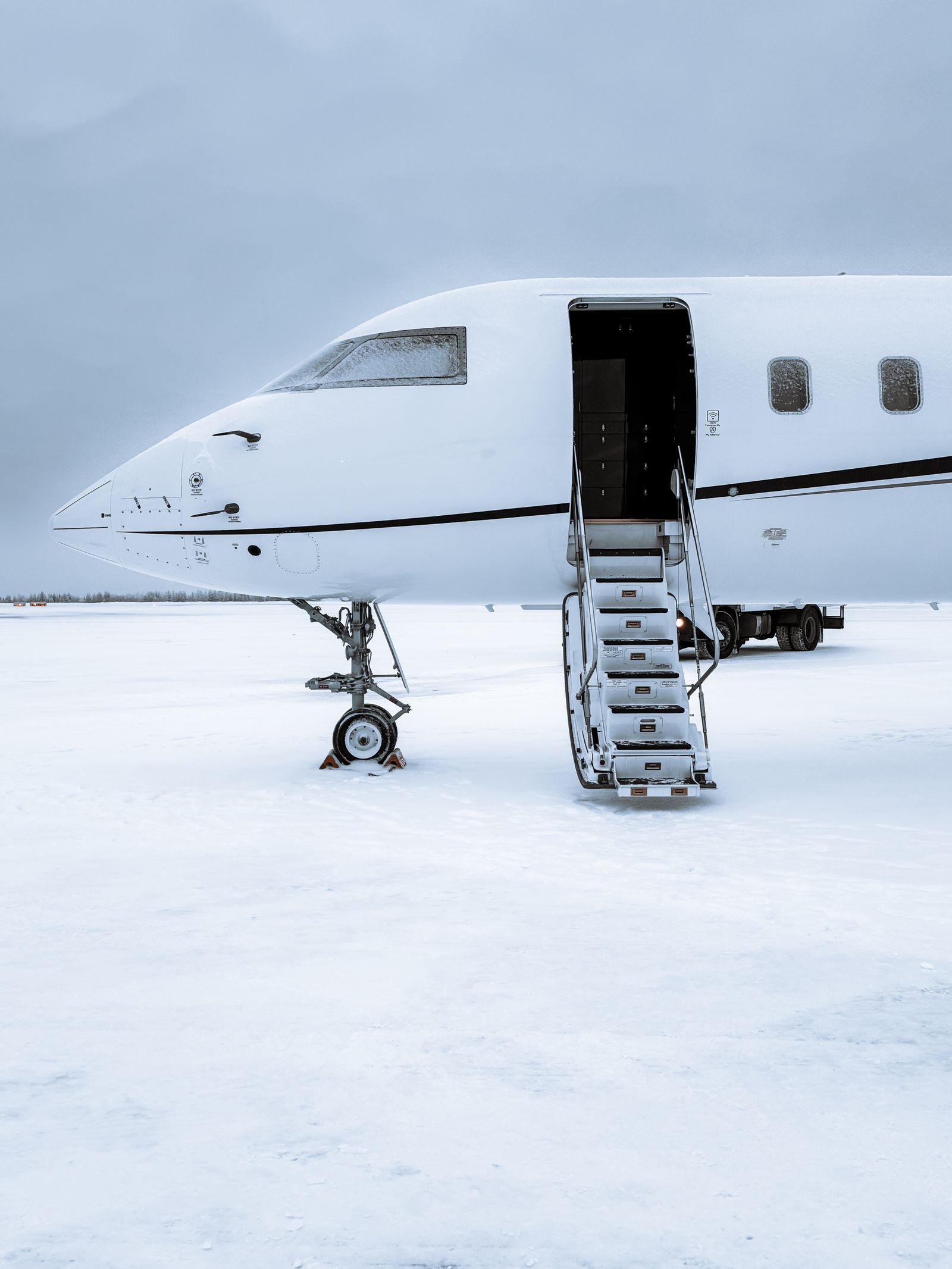 A white business jet parked on snow-covered ground with its airstairs lowered and its cabin door open, under a grey winter sky